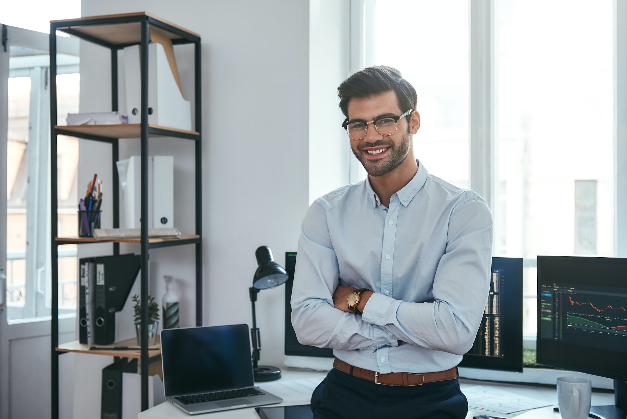 Happy trader. Cheerful businessman in formal clothes and eyeglasses is keeping arms crossed, smiling