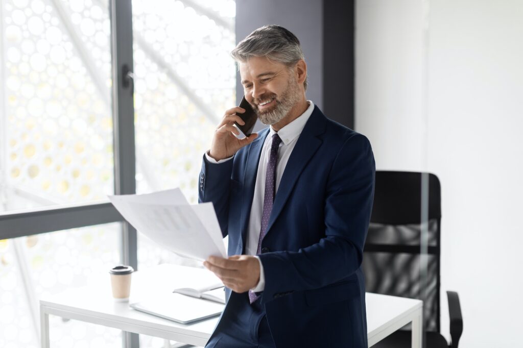 Smiling Middle Aged Businessman Talking On Cellphone And Checking Papers In Office